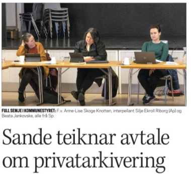 bilde frå eit forsamlingshus med scene i bakgrunnen. Tre kvinner sit ved sidan av kvarandre ved eit langbord med pcar framfor seg.